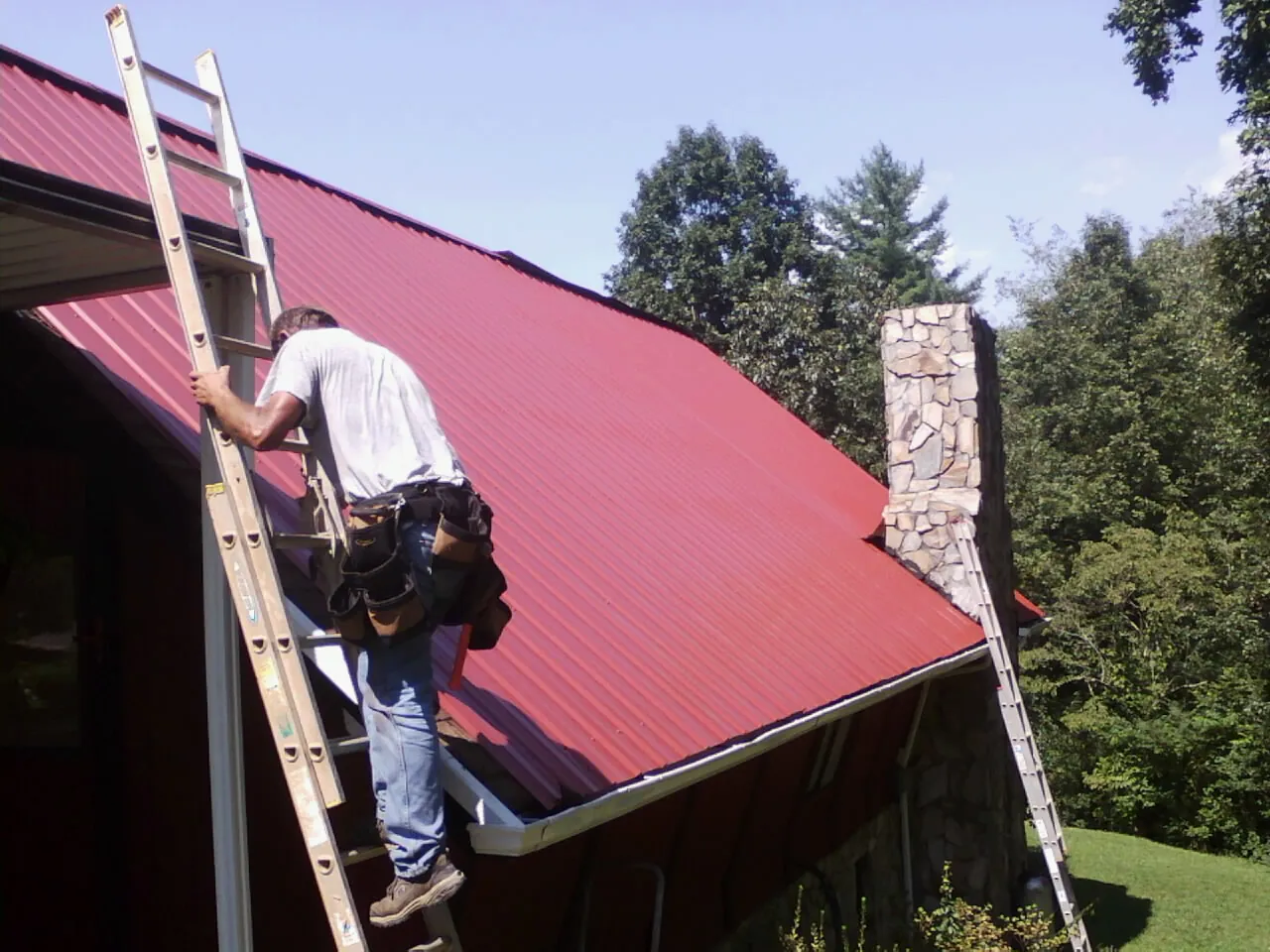Roofer on a ladder beside a red metal roof and stone chimney — Soffit & Fascia Repair by Roofing Brothers in Kearneysville